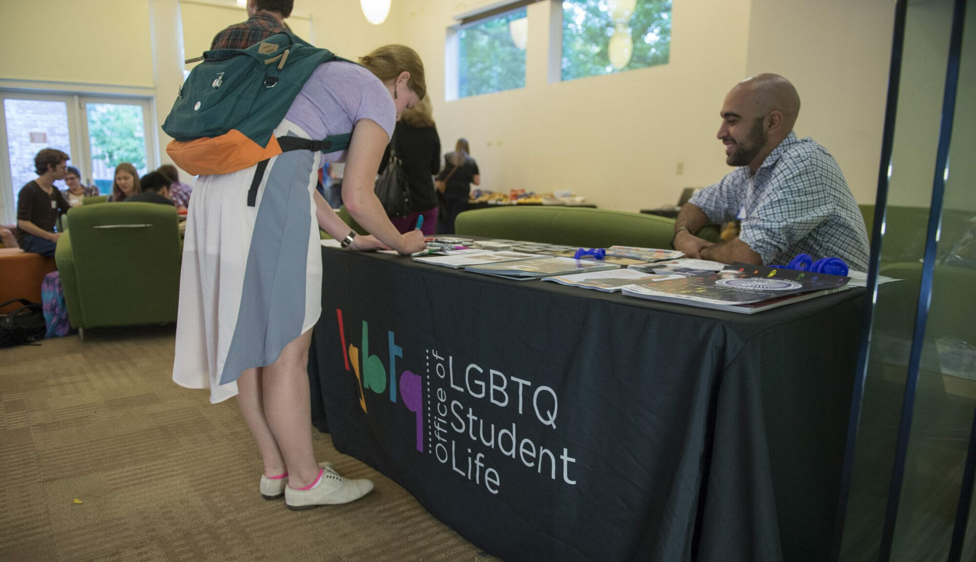 A student wearing a backpack writes a nametag at a check-in table with the LGBTQ Student Life logo