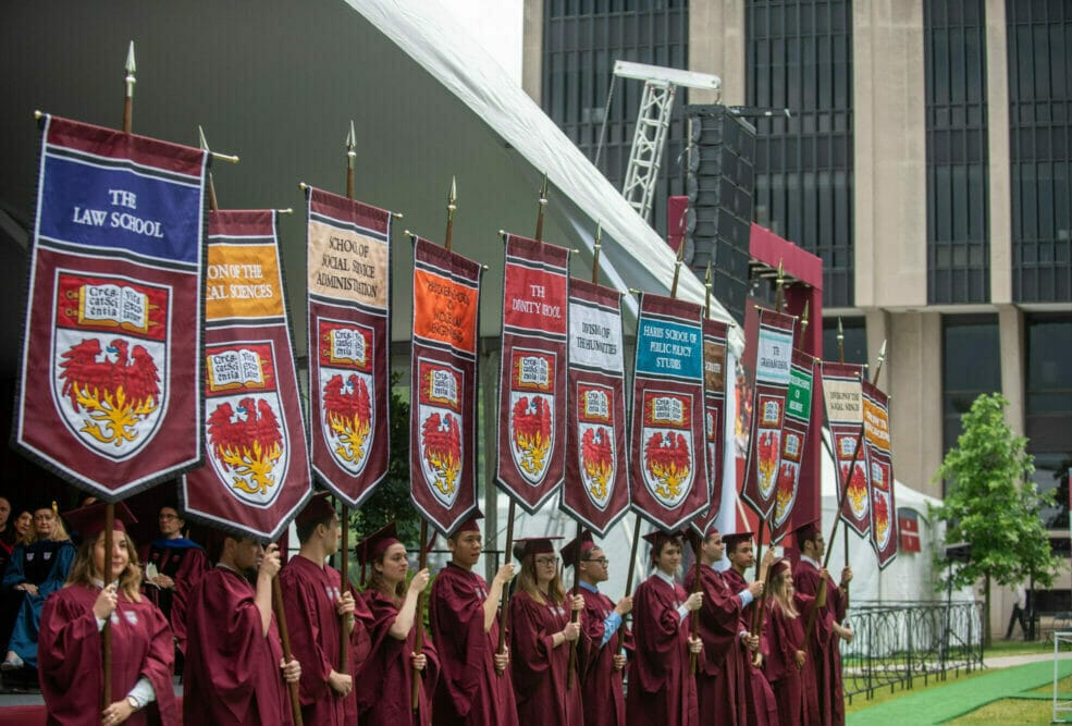 Students in graduation robes hold banners representing each graduate school